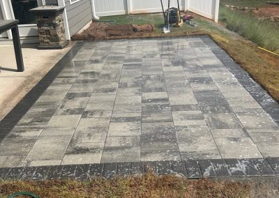 A newly installed gray stone patio sits beside a house, with tools and fresh soil around the edges.