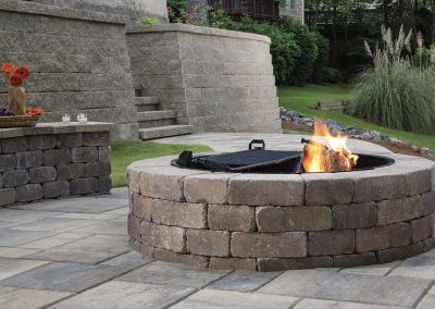 A stone fire pit with a small fire is set on a paved patio in a landscaped backyard.