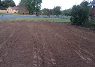 Freshly tilled dirt yard with visible tire tracks, bordered by grass, trees, and a brick house.