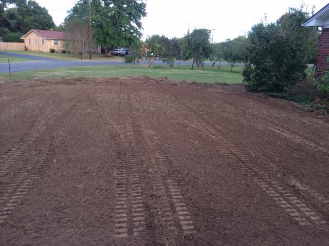 Freshly tilled dirt yard with visible tire tracks, bordered by grass, trees, and a brick house.