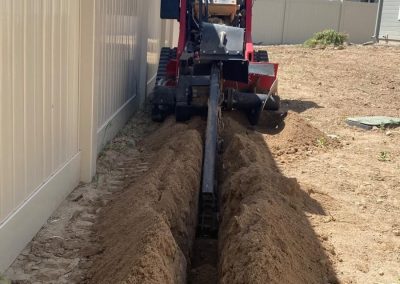 A small trencher machine digs a narrow trench next to a white fence in a backyard under a blue sky.