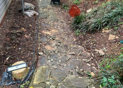 A stone pathway next to a house with a wheelbarrow, garden hose, and plants along the edges.