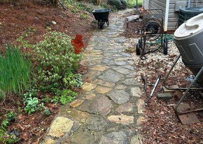 Stone path next to a house with a wheelbarrow, garden tools, and greenery on a rainy day.