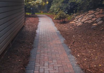 Brick pathway lined with mulch runs beside a house, surrounded by trees and greenery.