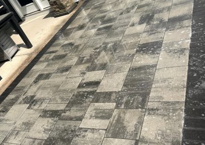 A backyard patio with a pattern of gray and beige rectangular pavers beside a house.