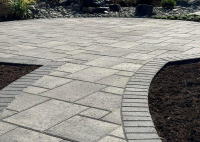 Curved stone patio with landscaped garden, shrubs, and a small rock fountain in the background.