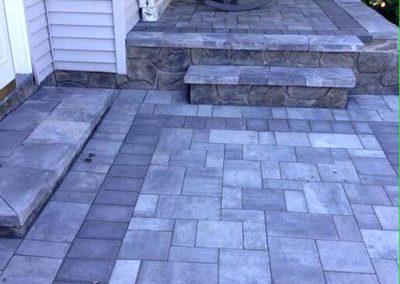 Front porch with gray stone pavers, steps, and a rocking chair next to a house with tan siding.