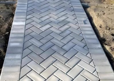 A paved walkway with a herringbone brick pattern bordered by rectangular bricks.