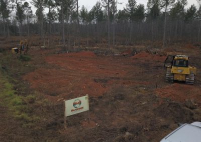 Two bulldozers clear red dirt at a forest edge; a sign and part of a car are visible in the foreground.