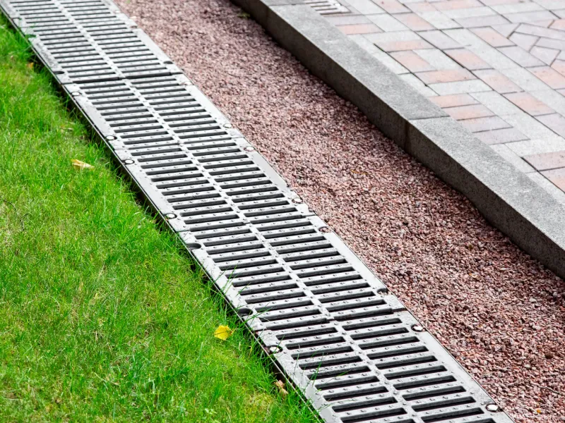 Metal drainage grate between green grass and a paved walkway with red gravel alongside it.