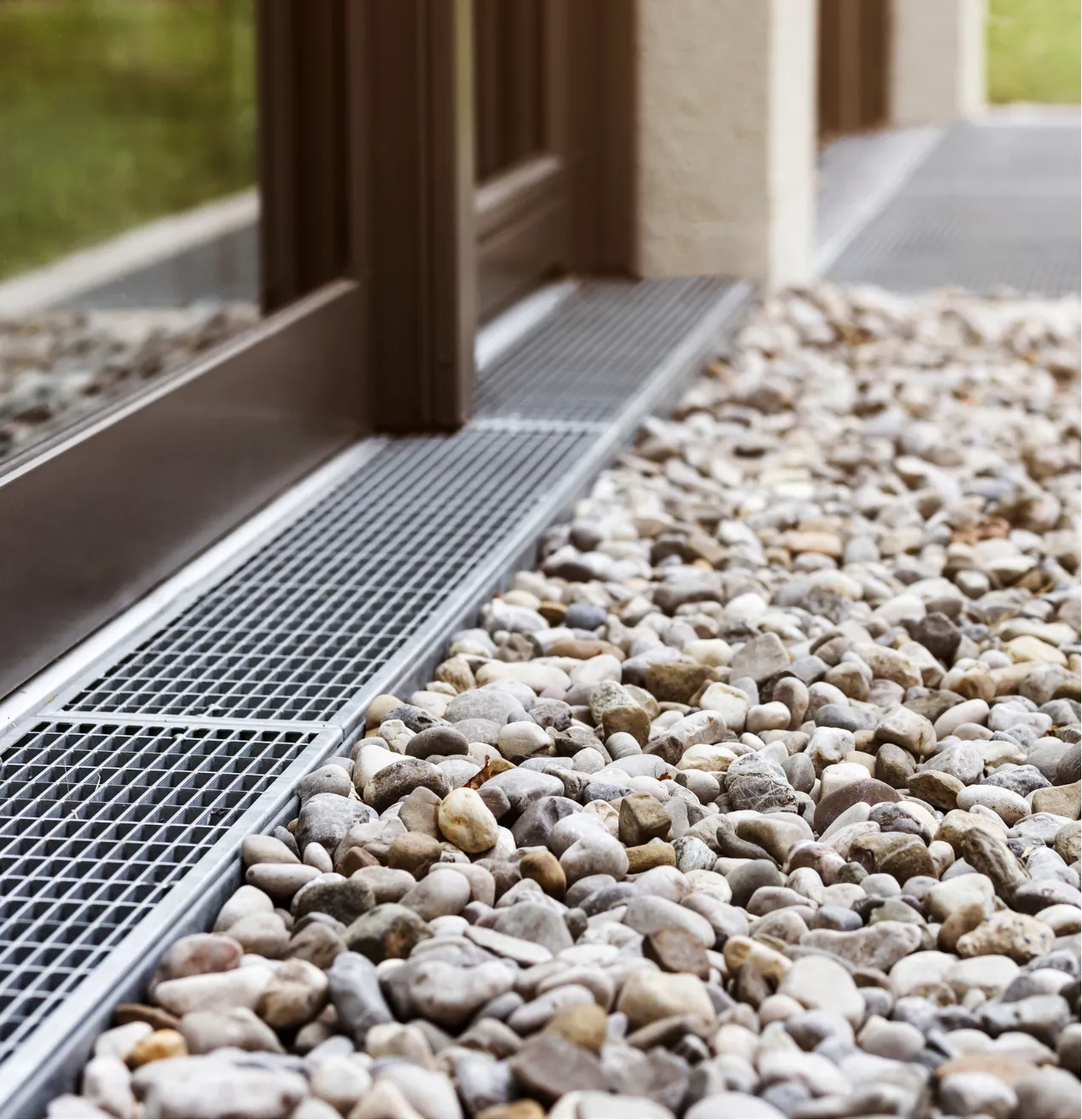 A metal drainage grate beside a building, surrounded by small stones and pebbles.